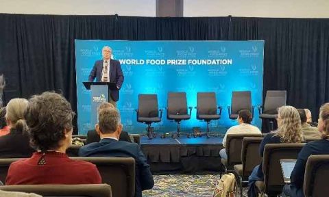 Man standing at lectern in front of "World Food Prize Foundation" sign, with four empty chairs stage right, audience in foreground