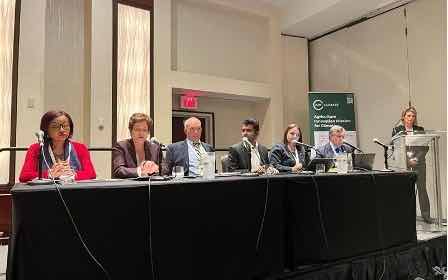 Six panelists seated at table; speaker at lectern on left