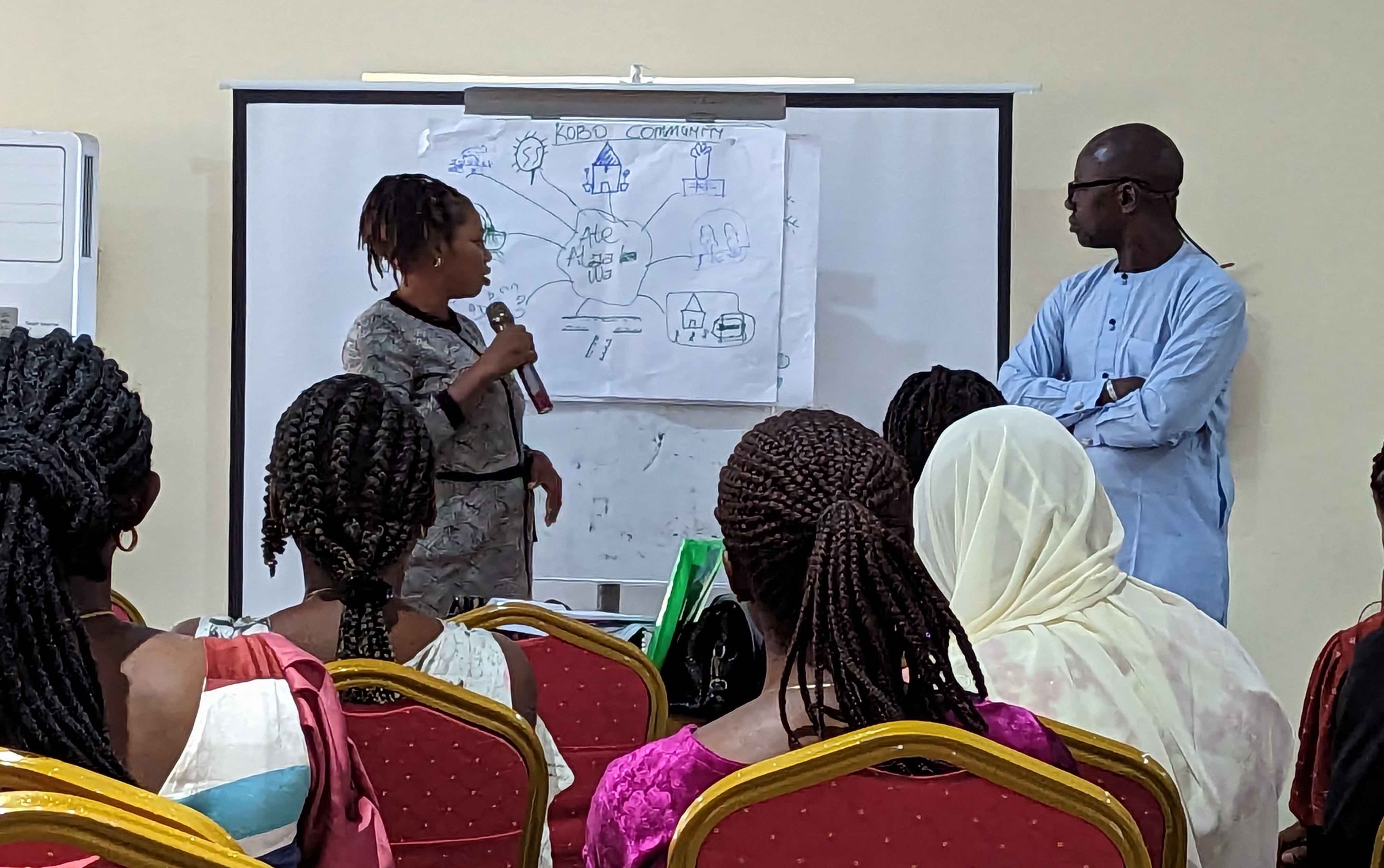 Woman holding microphone standing in front of whiteboard with drawing; man standing to right, audience with backs to camera