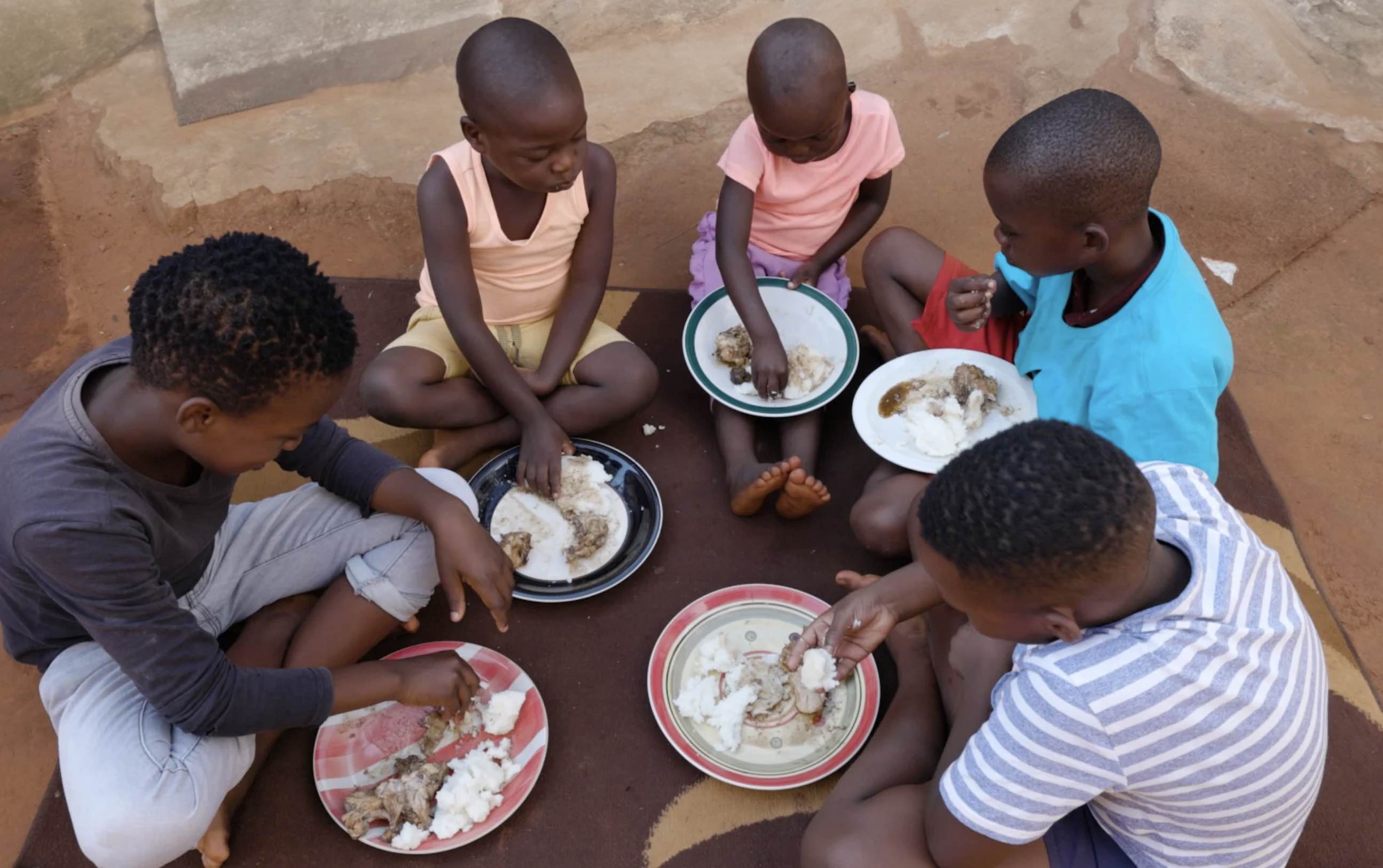 Five children seated in a circle on a rug eating