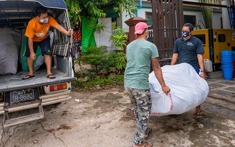 Man stands in truck bed, left, while two men carry big bag of hygiene kits into building