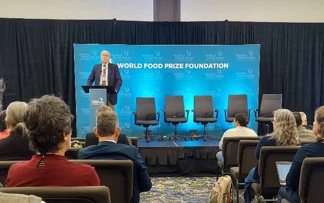 Man standing at lectern in front of "World Food Prize Foundation" sign, with four empty chairs stage right, audience in foreground