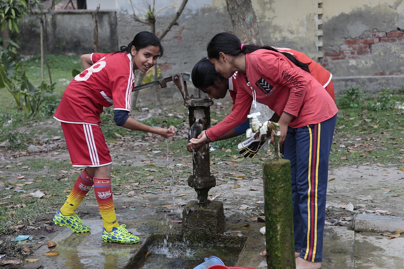 Three teen girls in athletic clothes wash hands at an outdoor pump