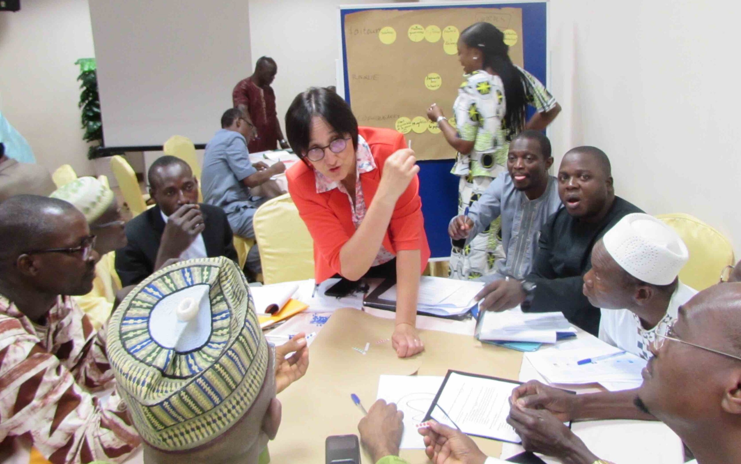 Woman, standing center at table, surrounded by seated training participants