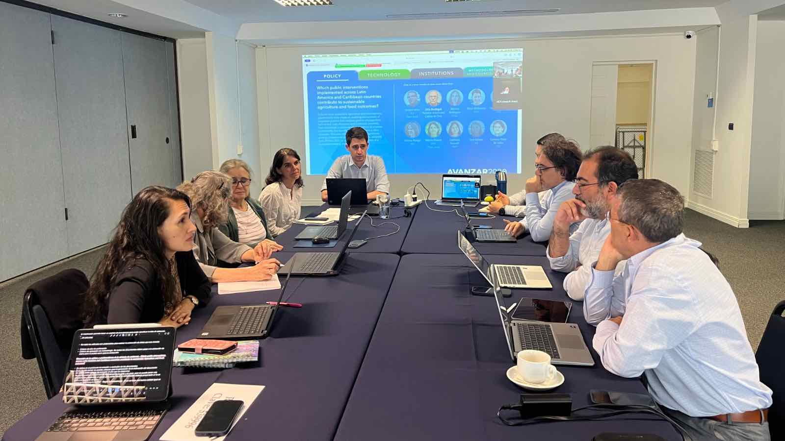 Workshop attendees seated on both sides of a table, with screen at rear