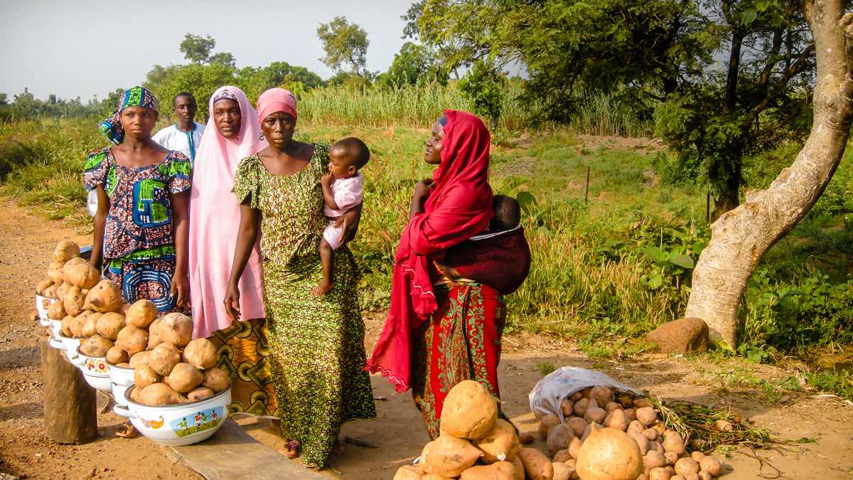 Four women, two carrying babies, with man in background, stand next to bowls of sweet potatoes lined up on a plank.
