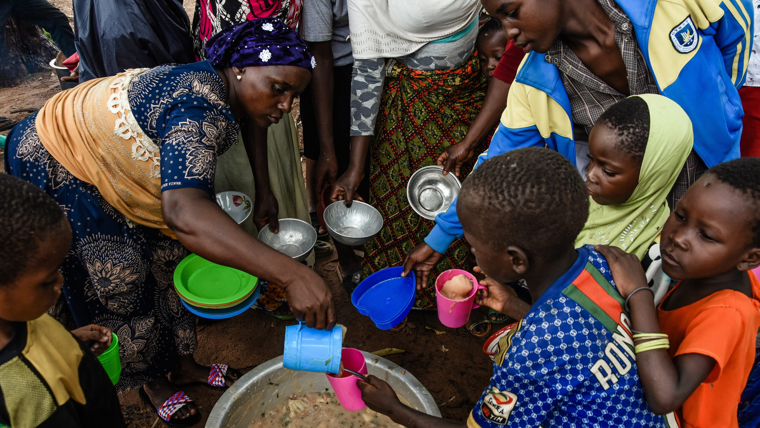 Woman, left, pours stew from a pot into a child's cup; other children and adults with cups/bowls surround them.