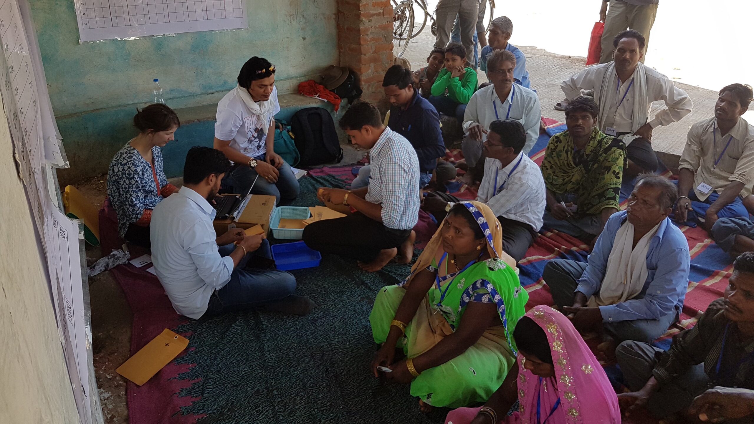 Men and women seated on a rug in shade, some in discussion, some looking at poster.