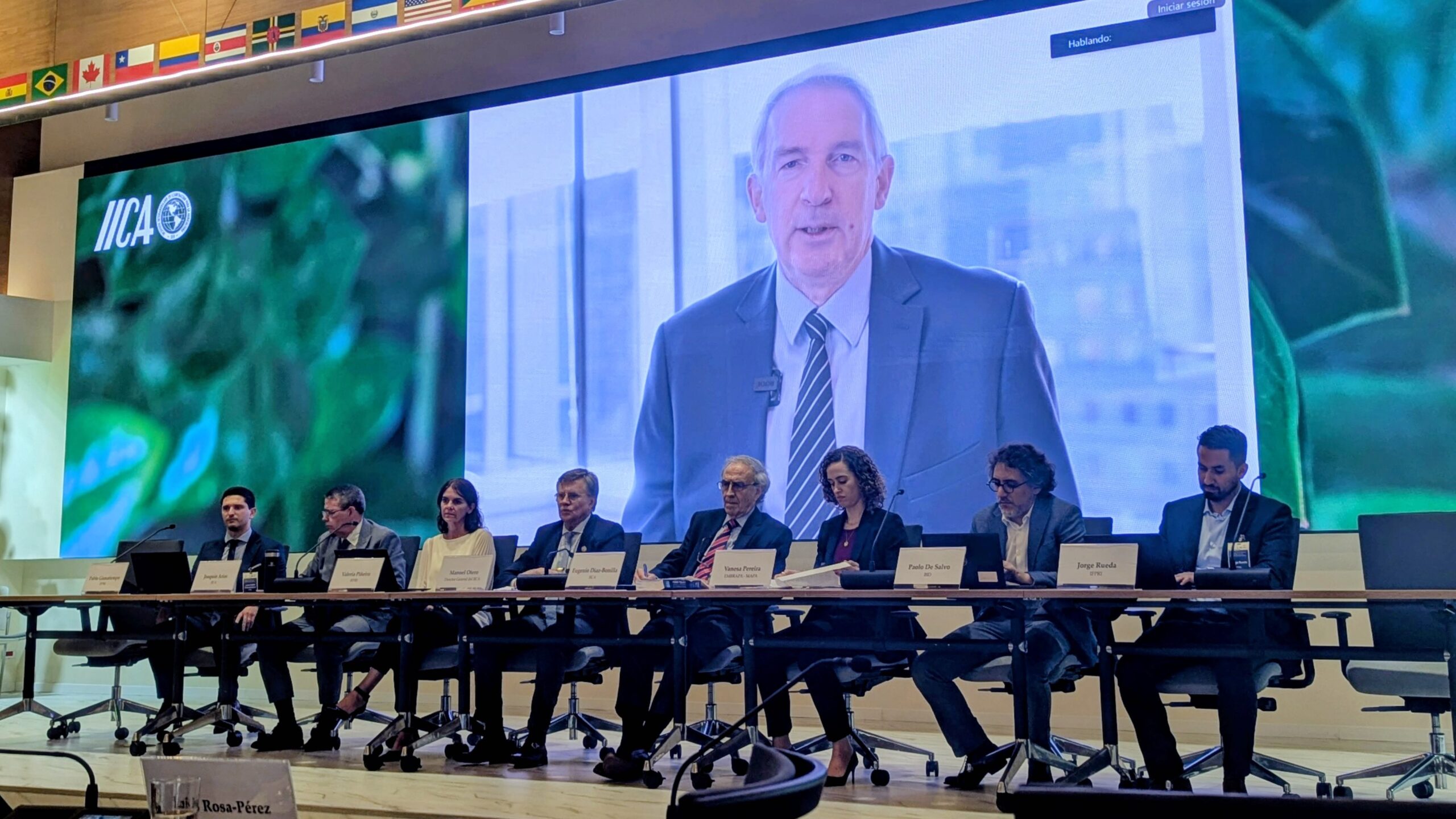 Panelists sit behind table on dais; image of man on screen behind them.