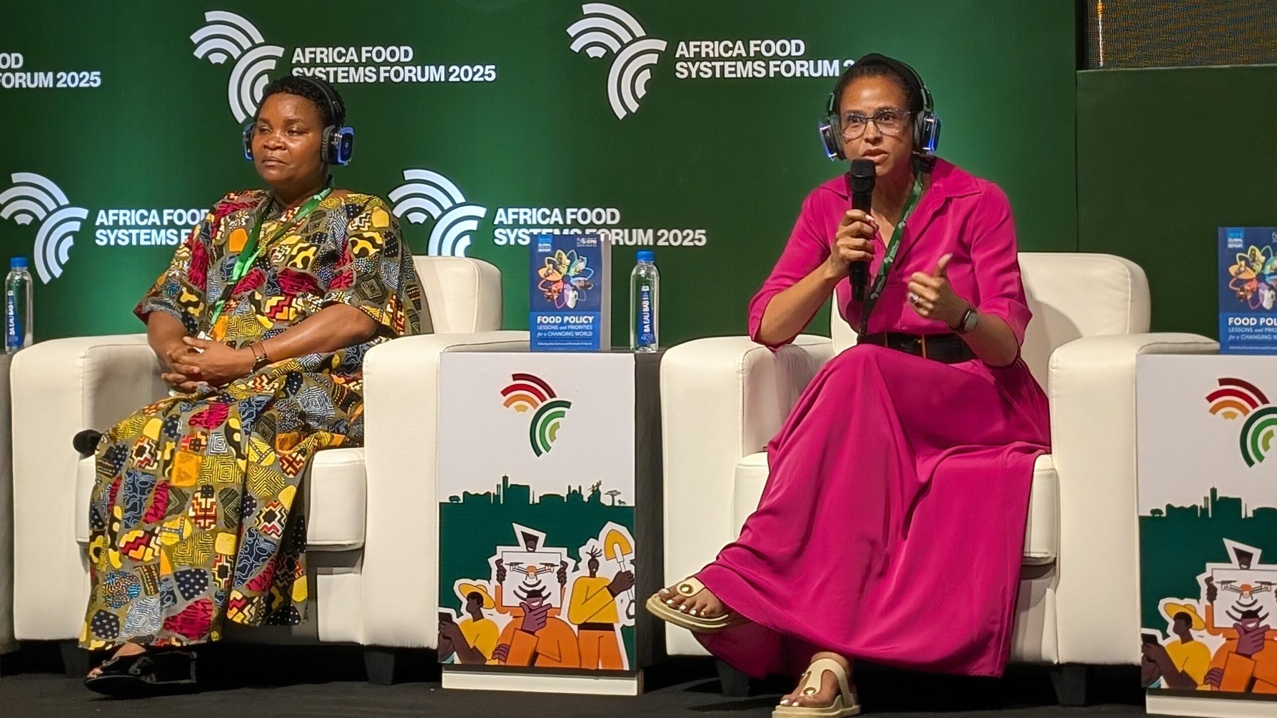 Two women seated in upholstered chairs on stage, woman on right holding microphone and speaking