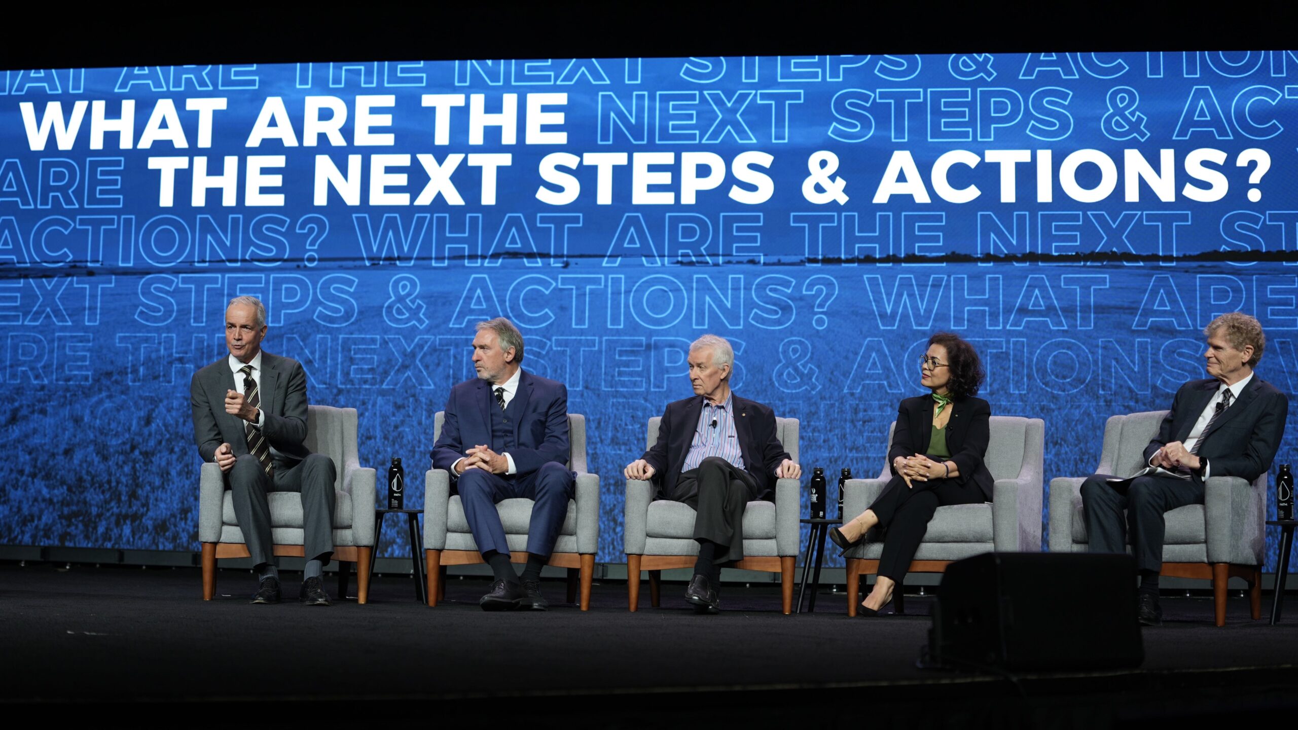 Five speakers sitting on stage under projected phrase: "WHAT ARE THE NEXT STEPS & ACTIONS"