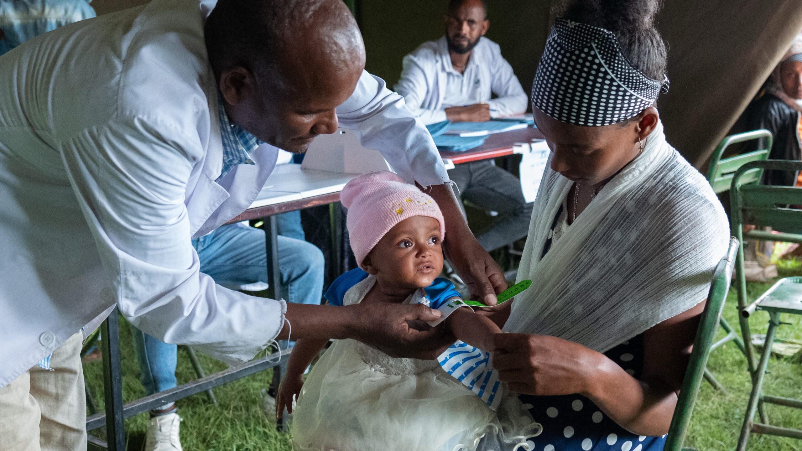 Man, left, wraps tape measure around army of baby, center, in arms of mother, right