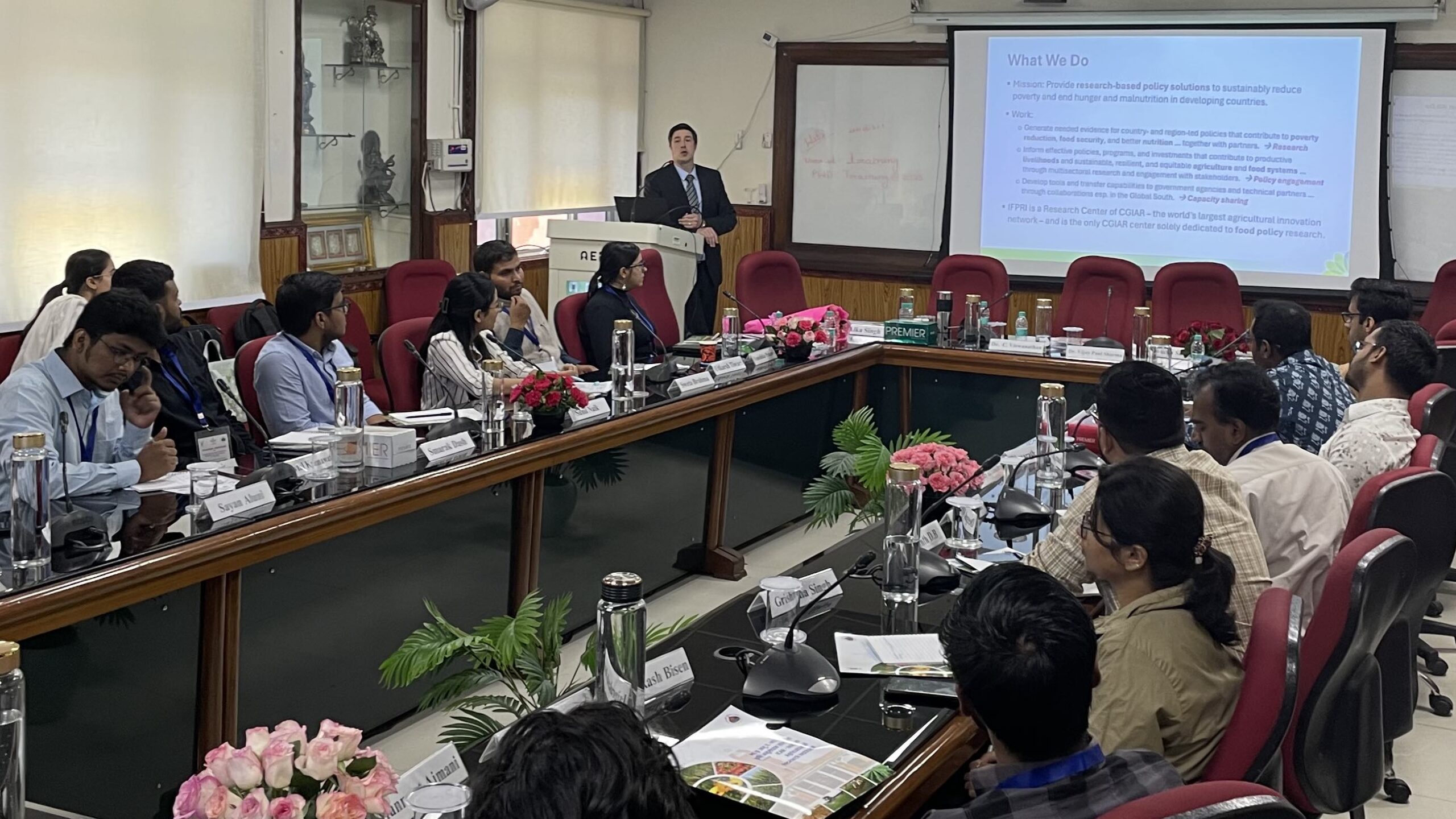 Man standing at lectern next to screen, center rear. Men and women seated around tables arranged in U shape, foreground.