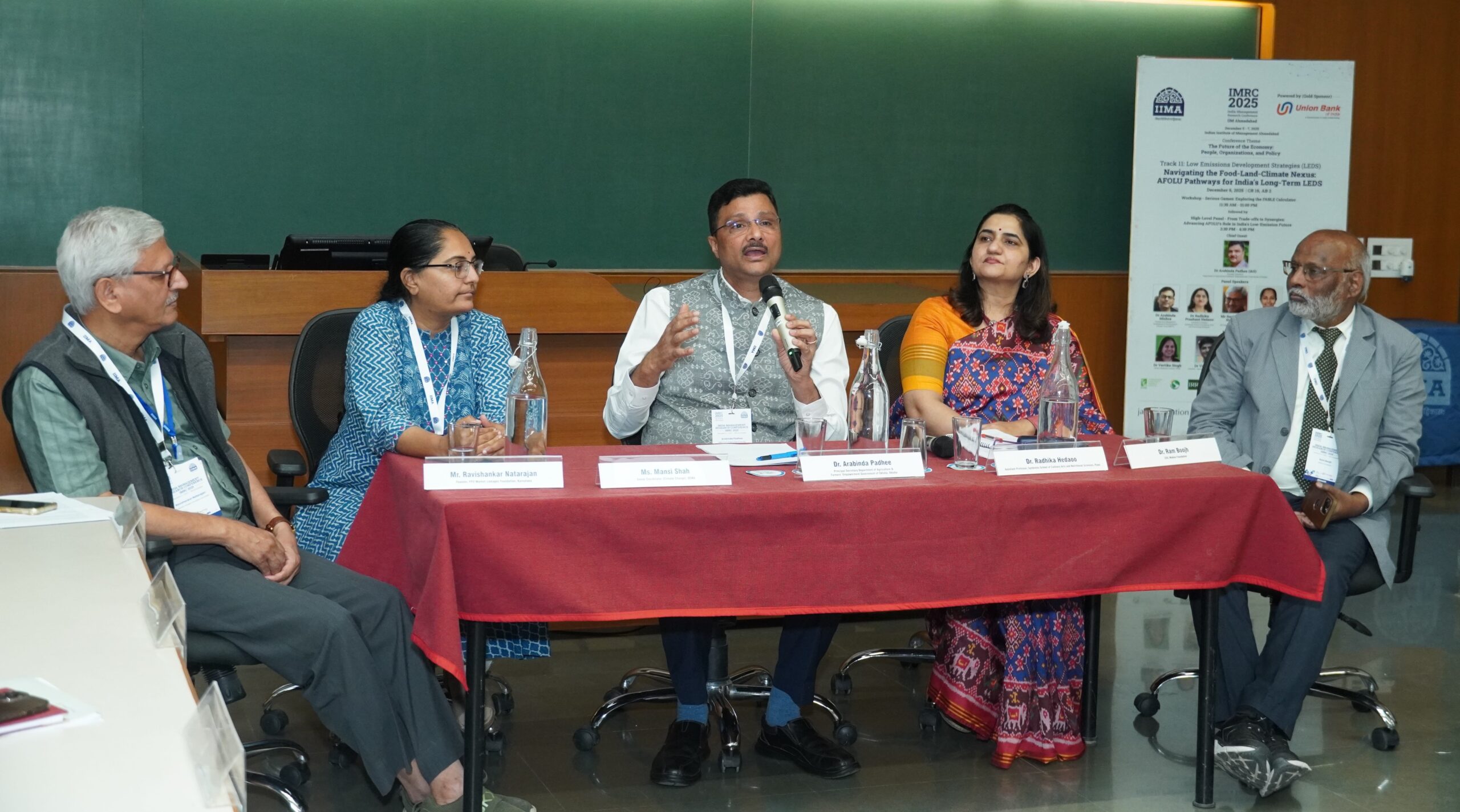 Five panelists sitting at table; man, center, holding microphone and speaking