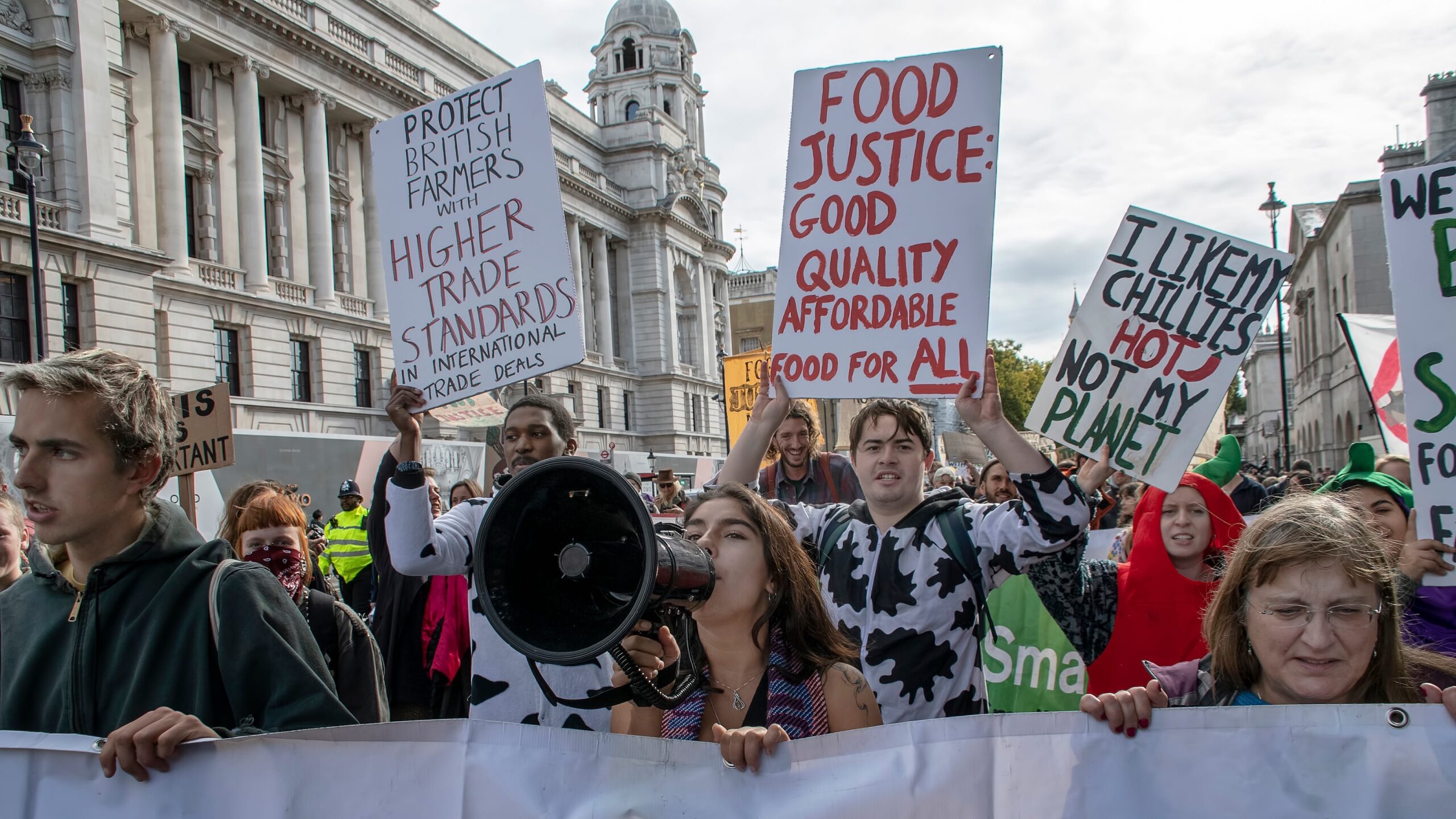 Protestors in street hoist signs including "Protect British Farmers with HIgher Trade Standards" and "Food Justice: Good Quality Affordable Food for All". Woman in center speaking through bullhorn.