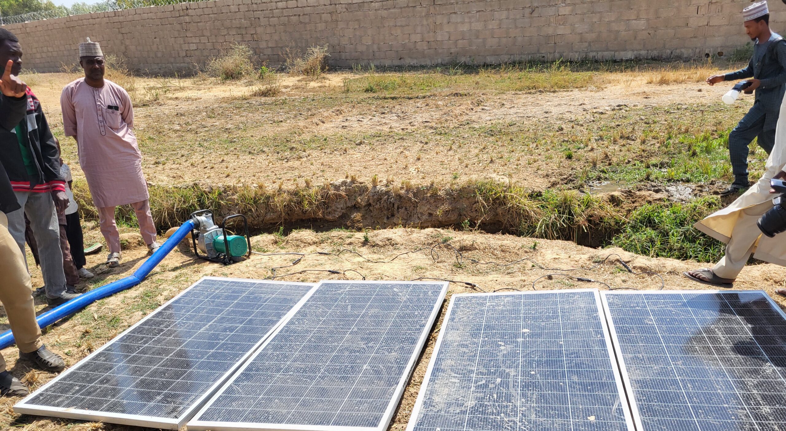 Men standing at far left and far right; pump, left, four solar panels flat on ground, center