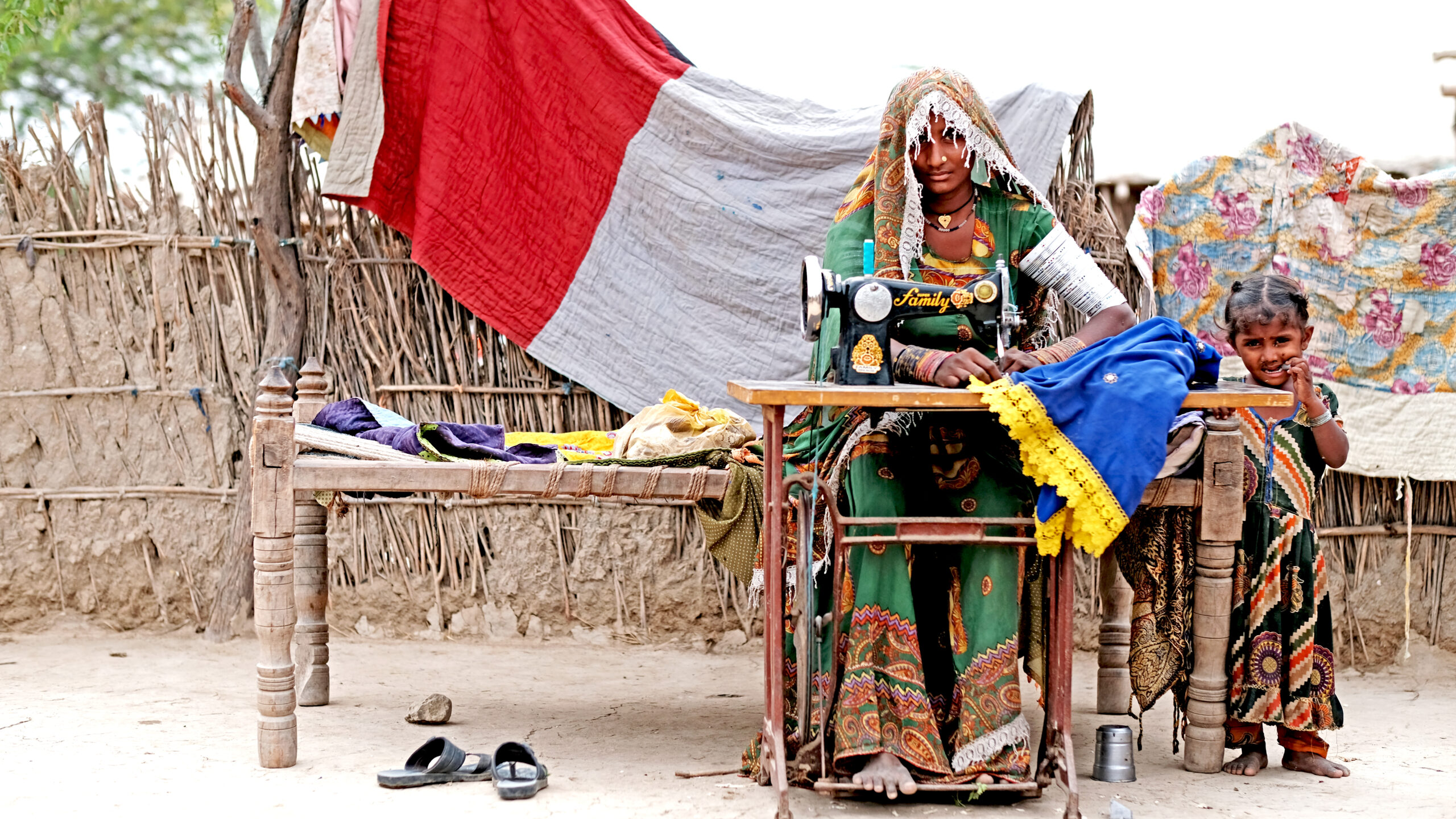 Woman sits at table outdoors operating sewing machine; child stands to right.