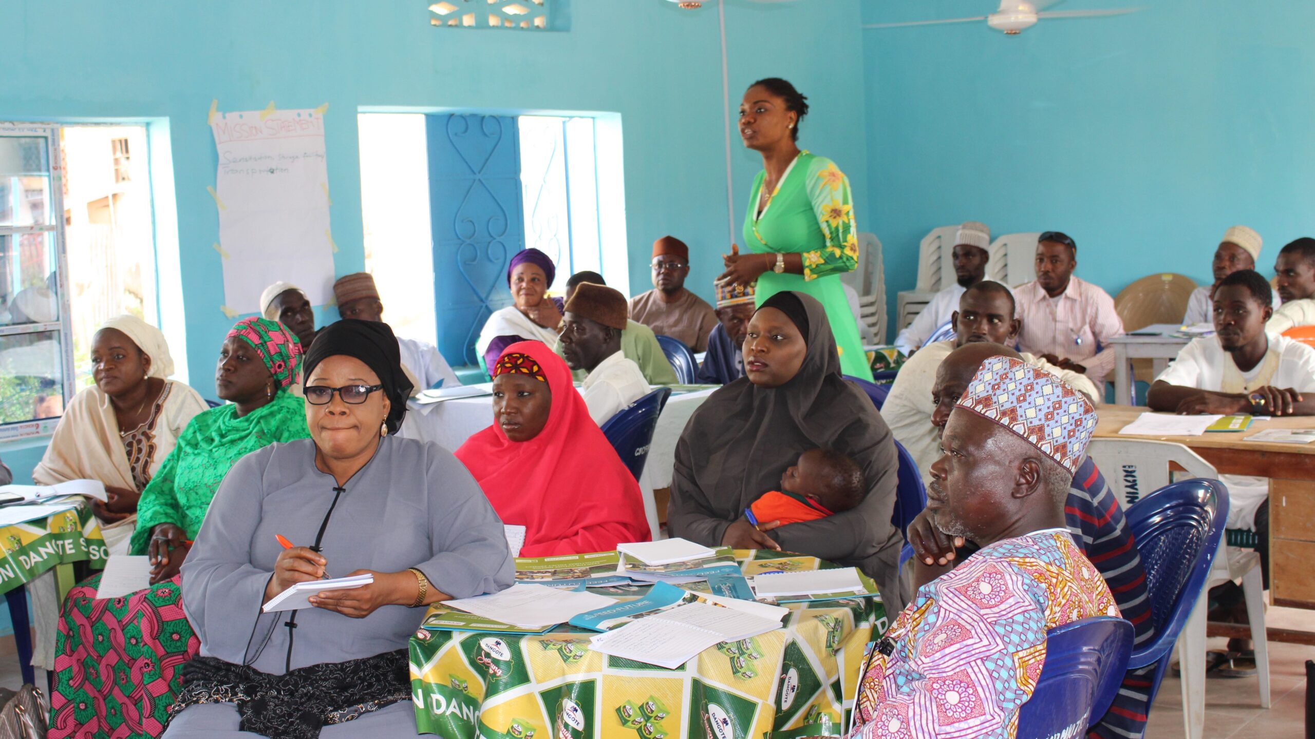 Men and women seated around tables facing left, listening. One woman, center rear, standing / speaking.