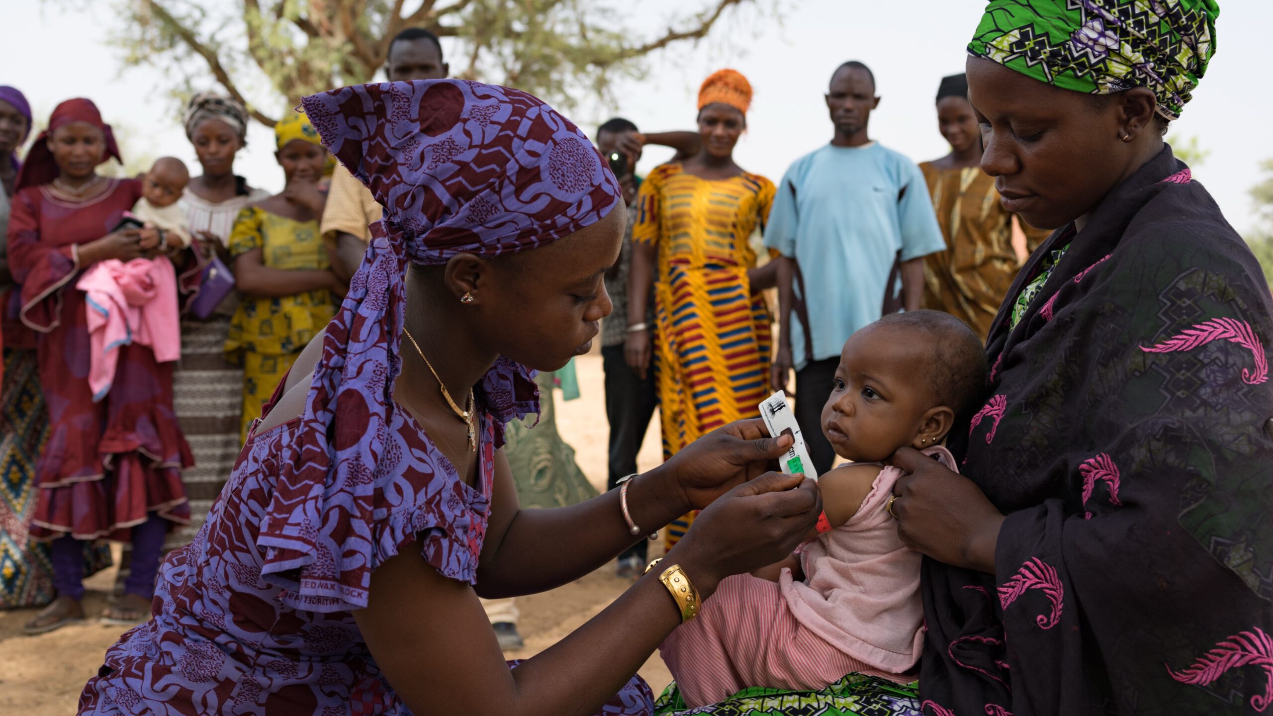 Woman, kneeling, left, measures baby's arm circumference. Baby is in arms of mother, right. Men and women stand in background.