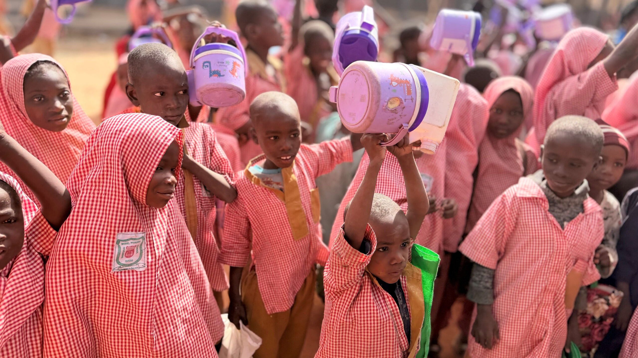 Children in red check school uniforms gathered outdoors, some holding up plastic food containers.
