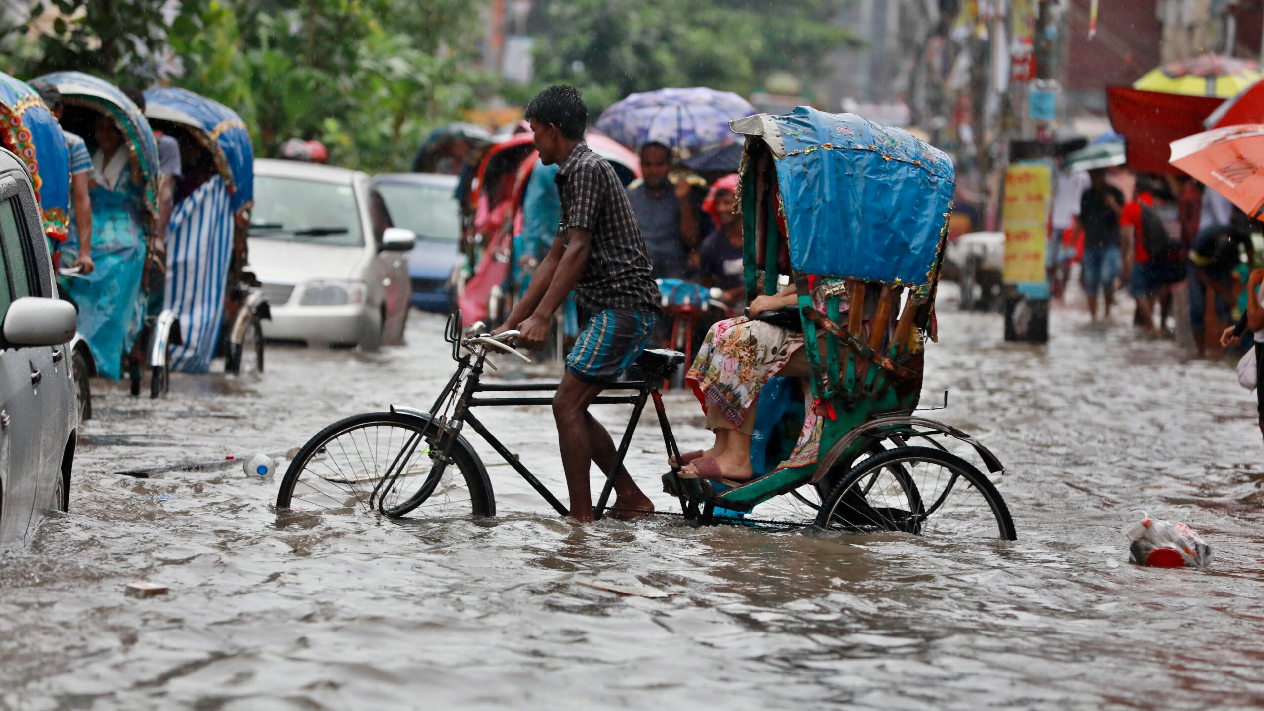 Man pedals bicycle with passengers in back through flooded urban street.