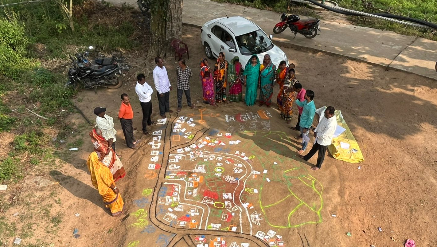 About 20 men and women stand in a circle around a drawing in the soil.