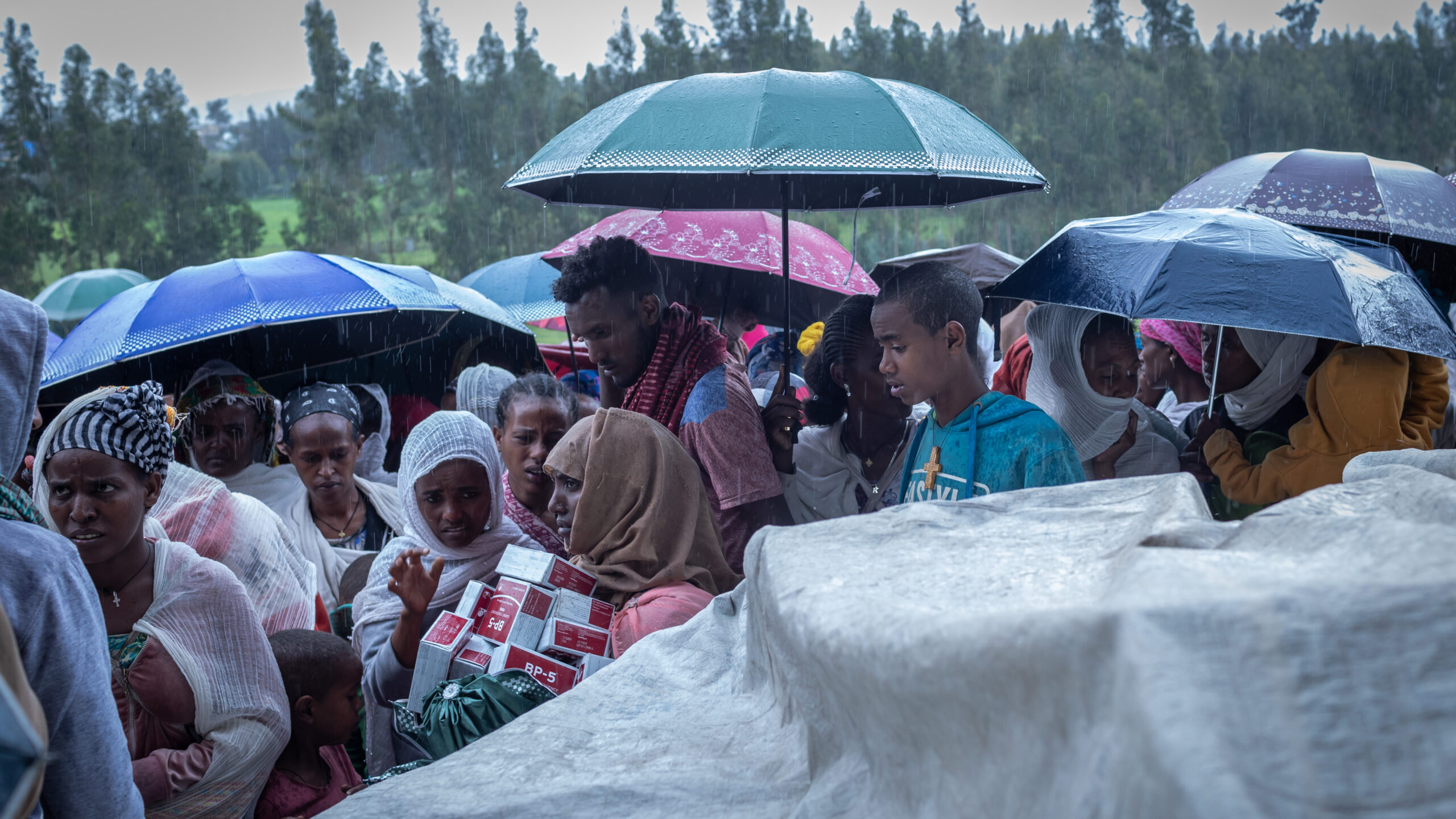 Men and women, some holding umbrellas, lined up; white tarp covered-object in right foreground.