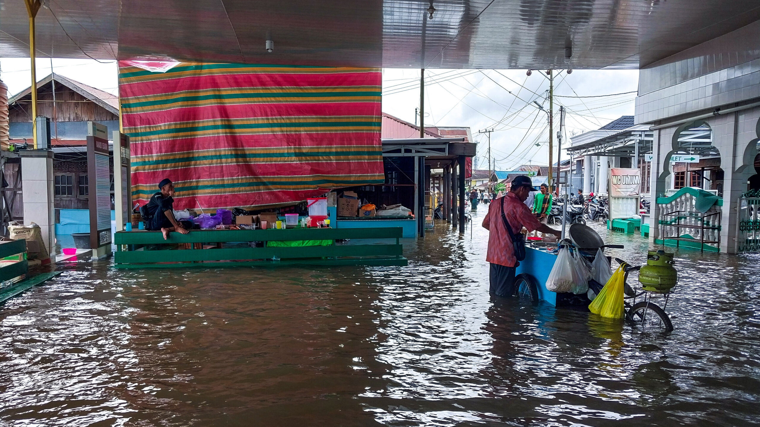 Flooded street market. Food / meal vendor on right. Man sitting on counter in background.