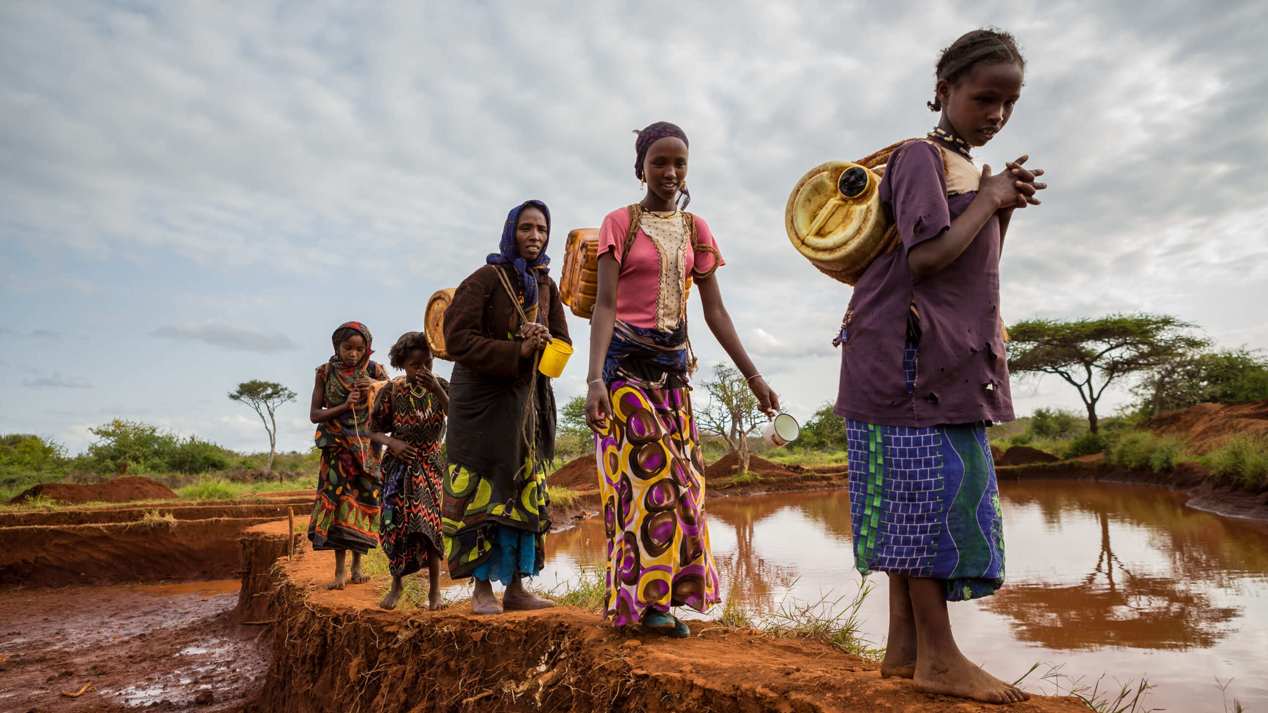 Five women walk along berm next to pond with containers.
