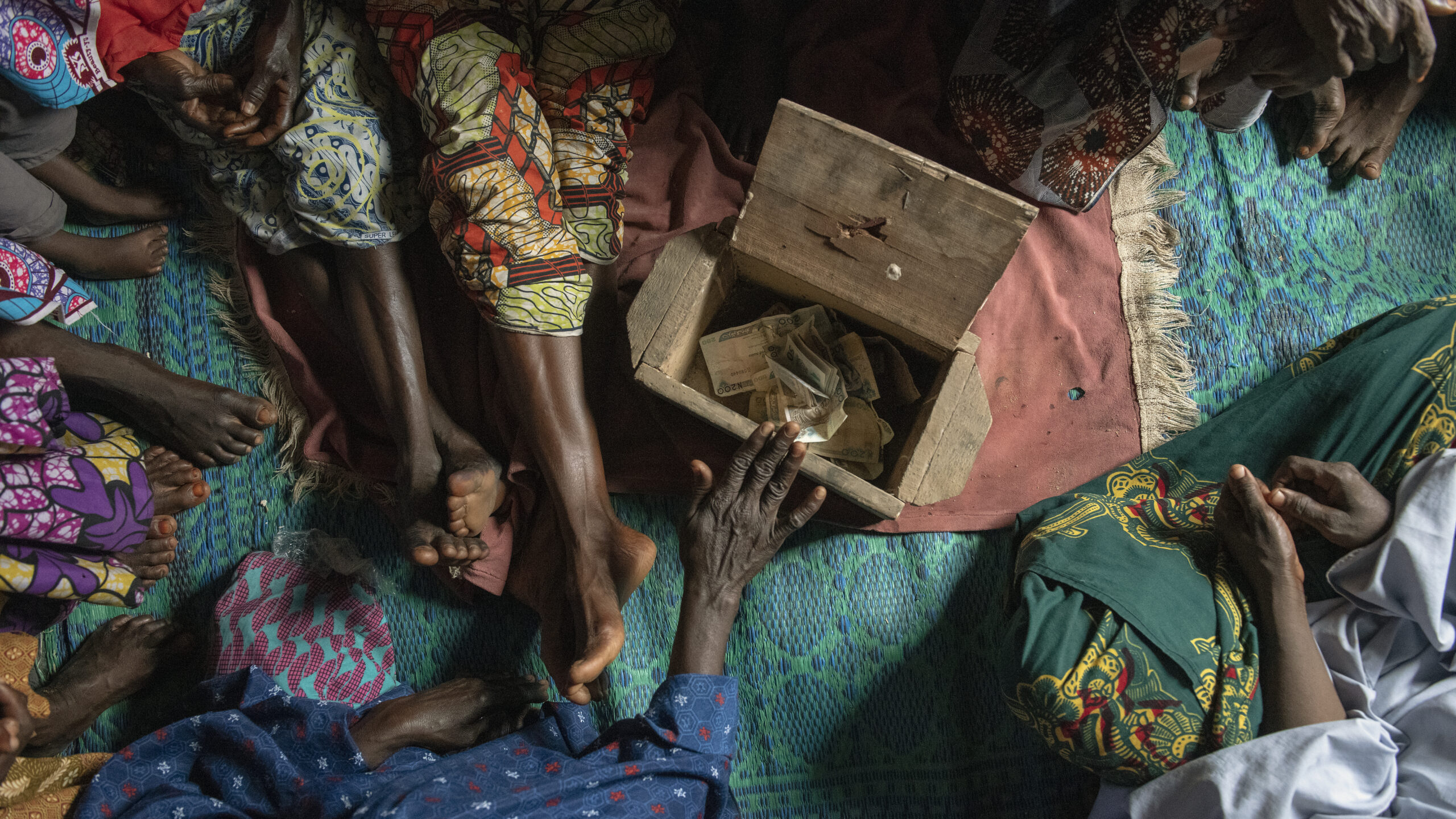 Hand on box holding money on blanket, center. Feet of women seated surrounding box in frame.