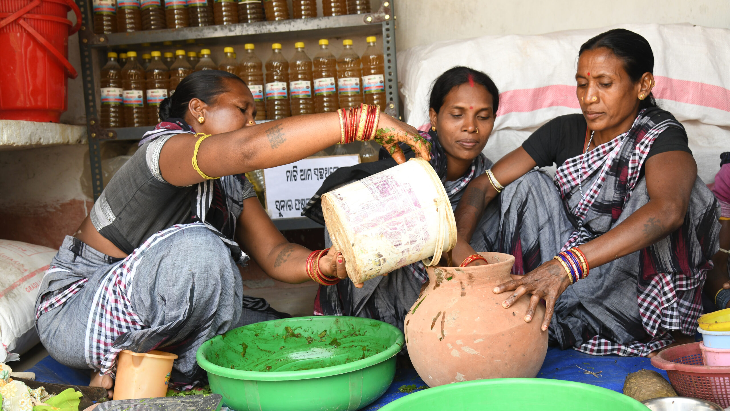 Three women seated on floor. One, left, pours bucket into bowl held by woman on right.