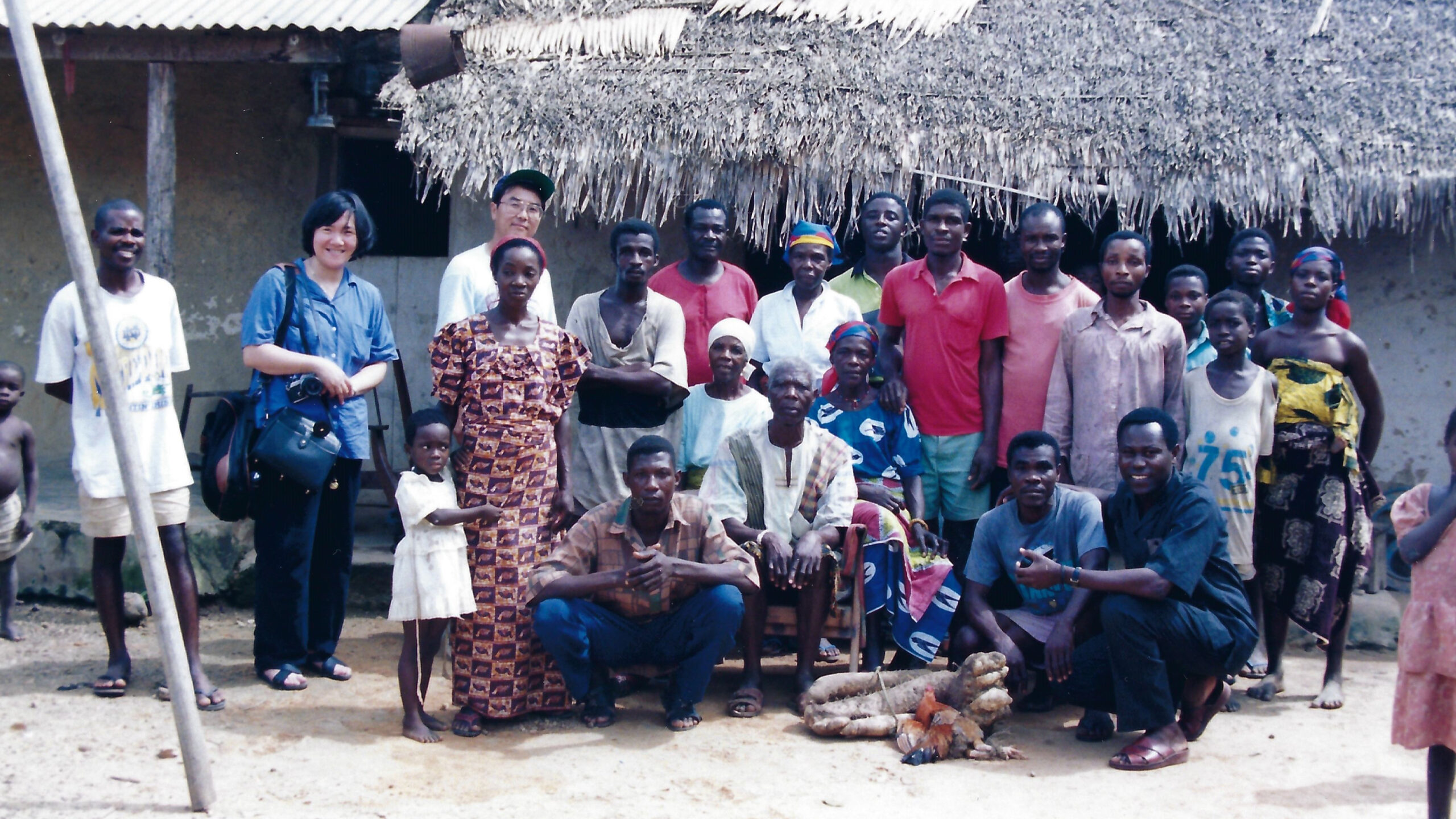 Woman and man, second and third from left, back row, standing with village residents, some standing some sitting on ground in front of building with corrugated metal and thatched roof.