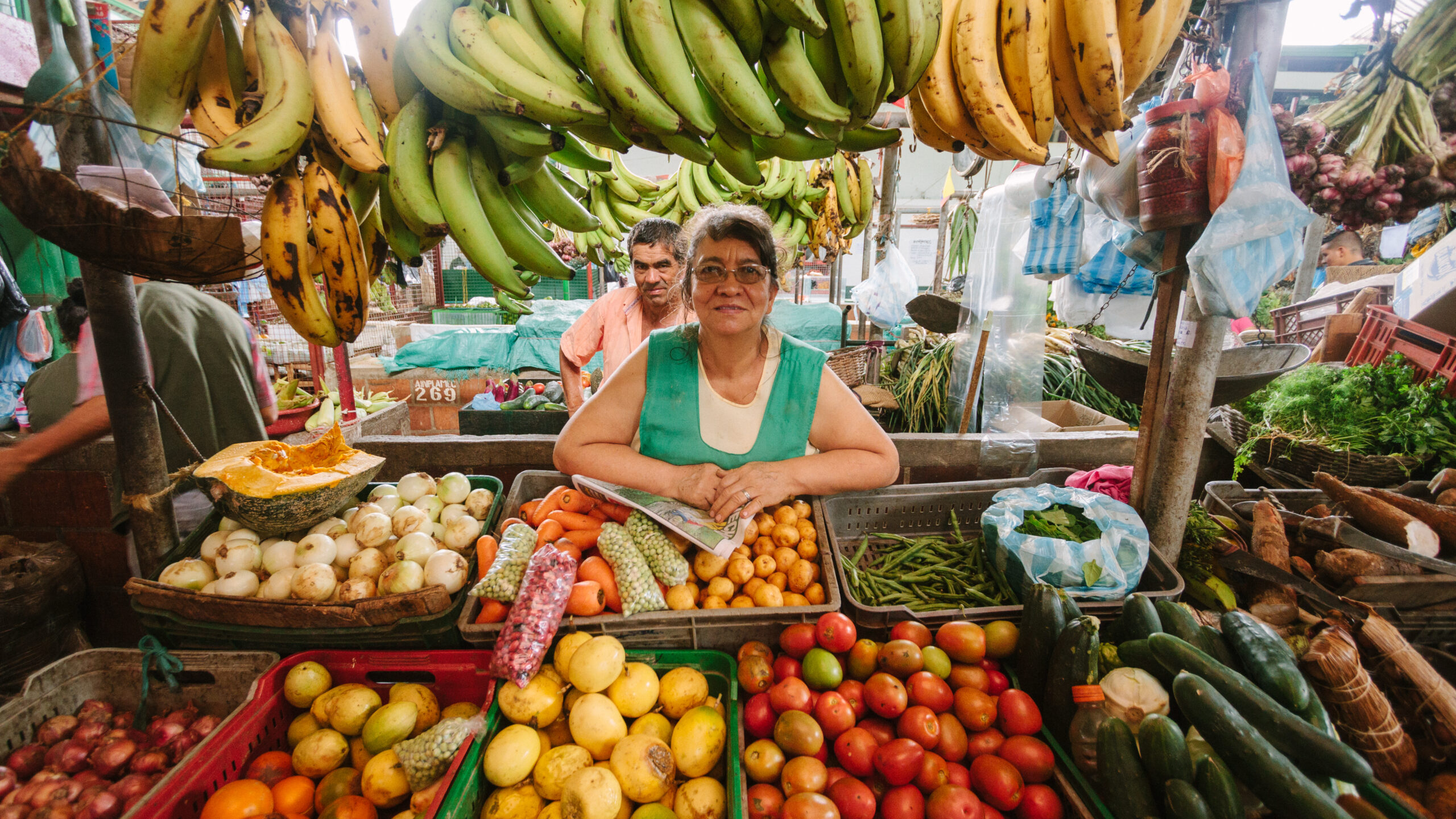 Woman and man behind her sit in a market stall surrounded by various kinds of produce including bananas, tomatoes, cucumbers