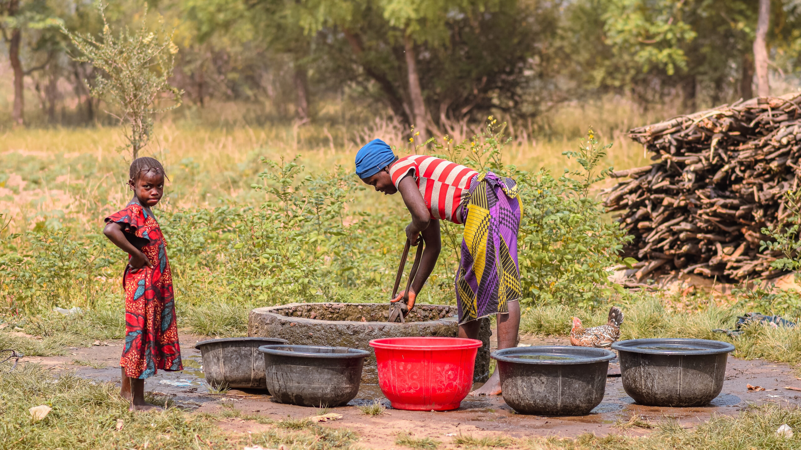 Girl, left, standing as woman, center, bends over pulling up water bucket from well. Five water containers on ground.