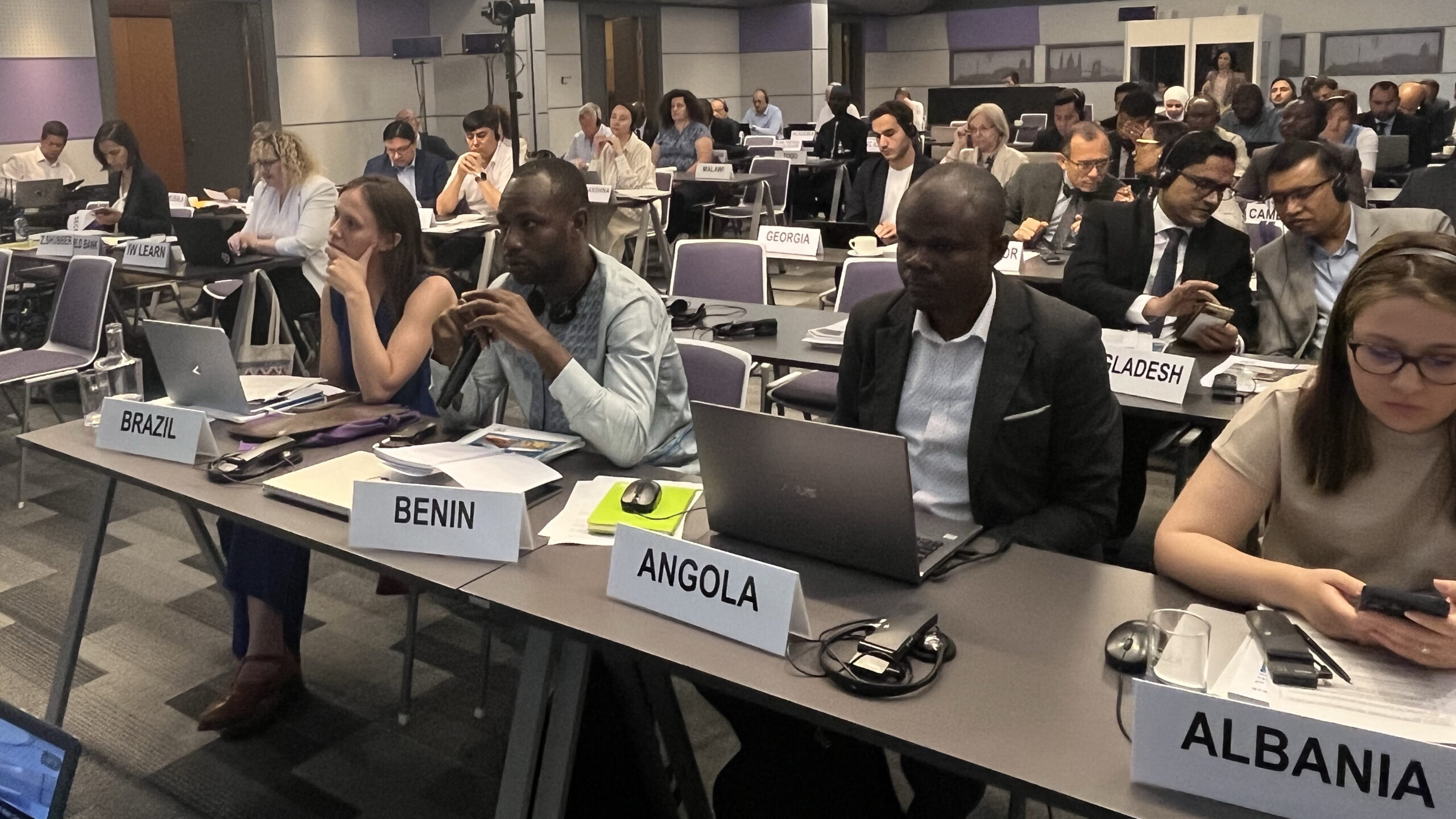 Men and women sitting at rows of tables in large room. Placards for people in first row read Benin, Brazil, Angola, Albania
