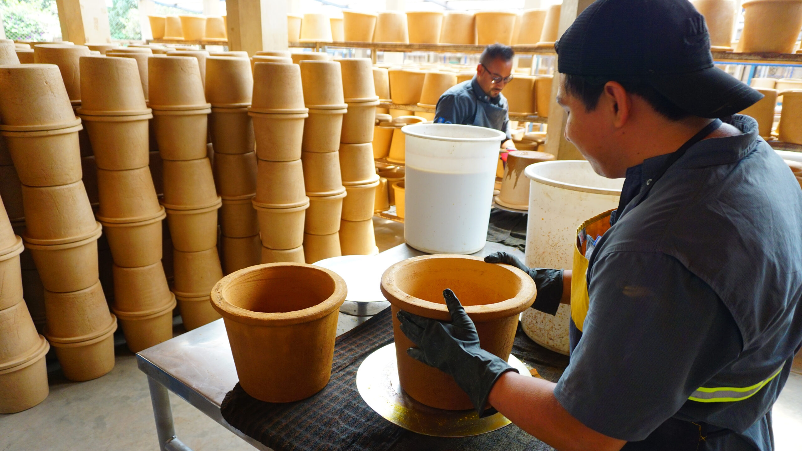 Man wearing baseball cap backwards, right, holds bucket filter; stacks of filters, left background; second man, center rear, looks down at bucket.