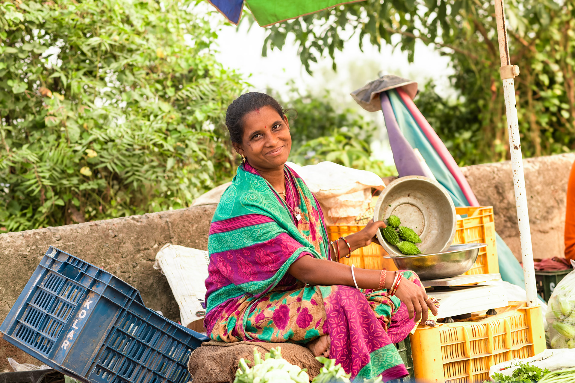 Woman in sari, seated, surrounded by crates, pours fruit from one bowl into another.