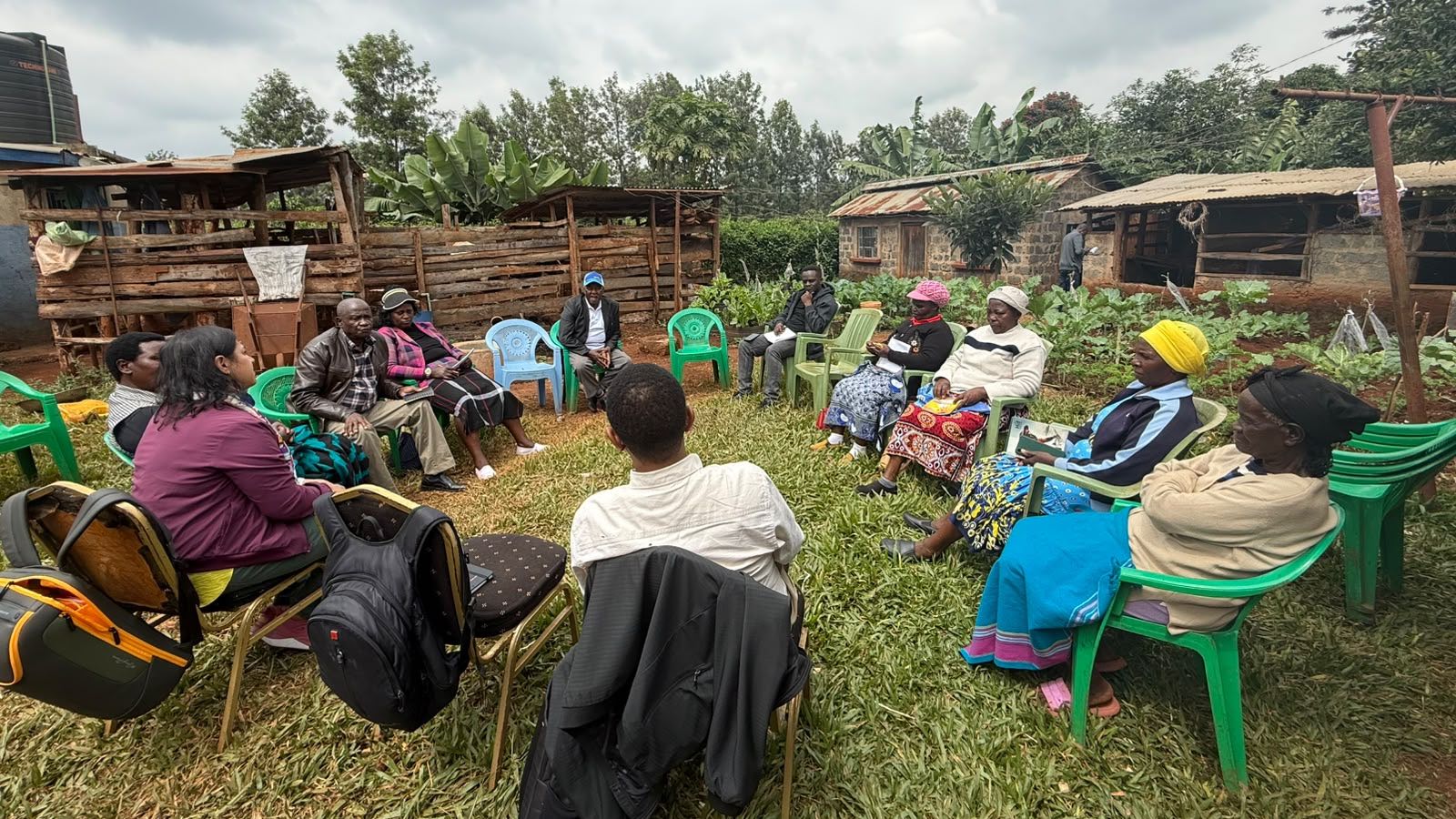 Men and women seated in chairs in a circle outdoors surrounded by livestock pens and other structures