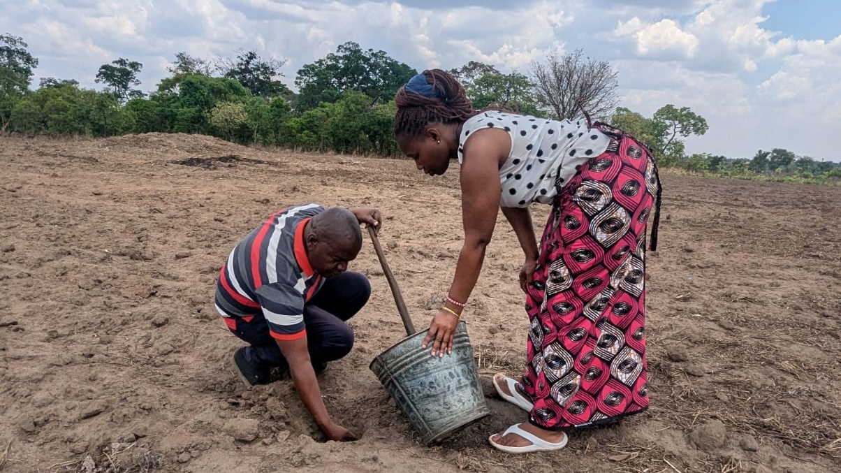 Man kneeling in field, right hand in soil, woman standing holding bucket.