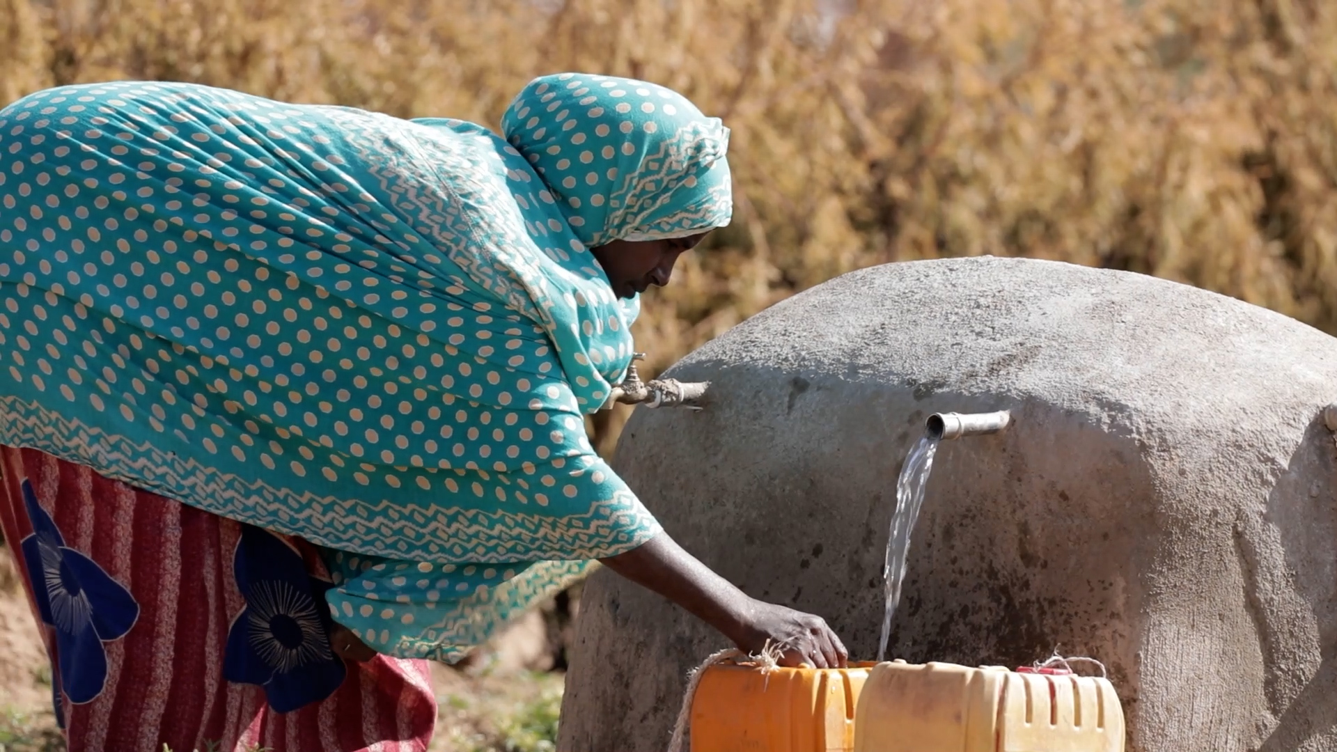 Woman left, bends over and puts hand in container as water flows into it from spigot.