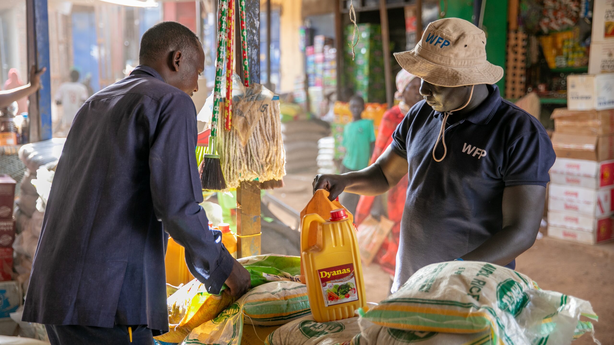 Man, left, facing right and grasping rice bag. Second man, right, wearing hat and shirt with WFP logo, grasping jug of fruit juice.